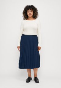 Marinblå plisserad kjol med svart midjeband, i kombination med en krämfärgad långärmad topp. Outfiten har släta texturer och en avslappnad passform.