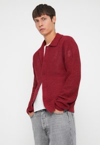 Red knitted cardigan with a button closure, featuring a textured pattern and a collar. Paired with a white shirt and gray jeans.