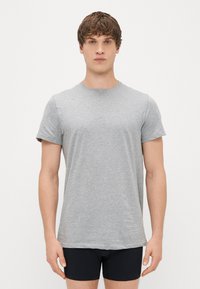 Pilkas trumpas rankovėmis marškinėliai iš minkštos medvilnės, su apvalia iškirpte ir laisva siluetu. Dėvimi su juodomis šortais, rodomi prieš baltą foną.