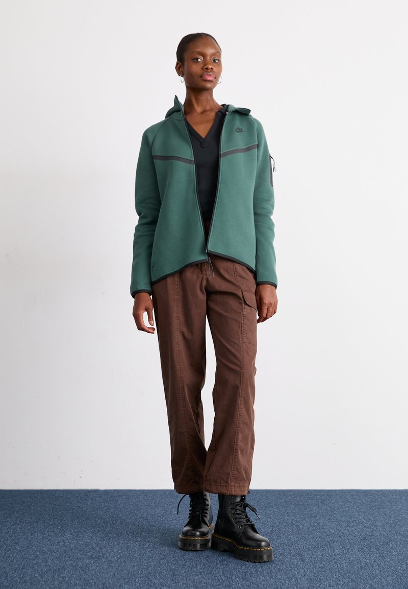 Haut à fermeture éclair vert avec un col montant, accents noirs et manches longues, associé à un pantalon cargo marron et des bottes noires à lacets.