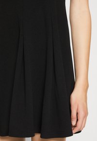 Robe plissée noire en tissu doux, avec un ourlet évasé et une texture lisse, avec un gros plan d'une main.