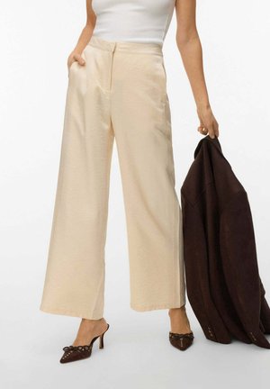 Donna che indossa pantaloni crema a gamba larga e tacchi a punta marroni, con in mano una giacca marrone scuro piegata nella mano destra.