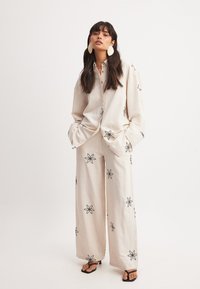 Béžový ľanový outfit s čiernym kvetinovým vyšívaním; voľný strih košele s golierom a široké nohavice; skombinované s čiernymi sandálmi na šnúrky.