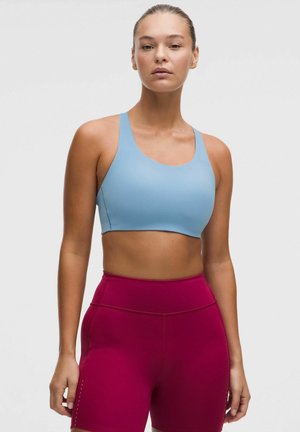 Vrouw staat, draagt een lichtblauwe sportbeha en hooggetailleerde magenta sportshorts tegen een effen grijze achtergrond.