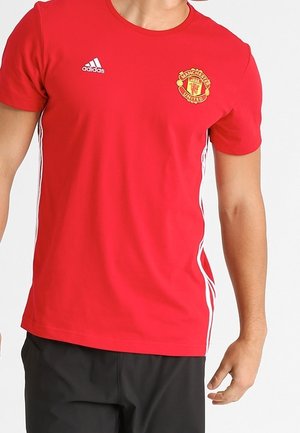 Czerwona koszulka Adidas Manchester United z białymi paskami po bokach, noszona przez osobę w krótkim rękawie i czarnych spodniach.