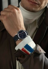 Zegarek chronograf z granatową tarczą, srebrnymi akcentami i teksturowanym niebieskim skórzanym paskiem. Noszony na białym żebrowanym golfie i brązowej kurtce.