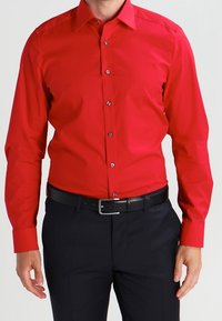 Rotes Langarmhemd aus Baumwolle mit glatter Textur; verfügt über eine durchgehende Knopfleiste und einen spitzen Kragen, getragen mit dunkler Hose und schwarzem Gürtel.