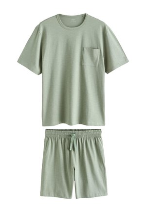 Person mit rechter Hand in der Tasche, trägt hellgrüne Freizeitshorts und ein passendes grünes Hemd vor neutralem Hintergrund.
