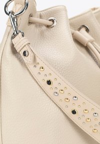 Sac en cuir texturé beige avec une sangle décorative ornée de clous dorés, argentés et noirs. Le matériel comprend un anneau métallique rond et un fermoir.