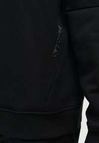 Κοντινή εικόνα μαύρου υφάσματος που δείχνει κάθετο λογότυπο Calvin Klein κοντά στη ραφή τσέπης σε ρούχο με μακρύ μανίκι.