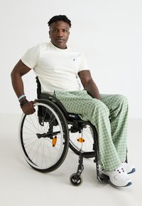 Fekete manuális kerekesszék szürke és fehér kerekekkel. Személy krém színű pólóban és zöld kockás nadrágban. Fehér sportcipő piros részletekkel.