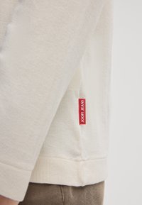 Helles beige Langarmhemd aus strukturiertem Stoff, mit einem roten "JOOP! JEANS"-Label an der Seitennaht. Der Manschette ist umgeschlagen.