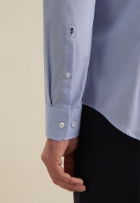 Camicia azzurra chiara con tessuto testurizzato, polsini abbottonati e dettaglio di un fiore ricamato sulla manica. Sono presenti bottoni decorativi.