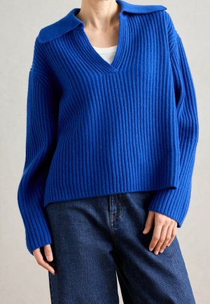 Person iført en knalblå ribstrikket sweater med krave og V-hals over en hvid top, kombineret med mørkeblå jeans.
