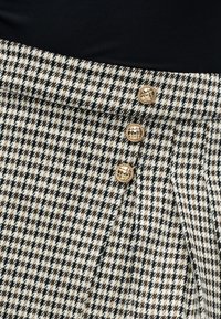 Φούστα σε σχέδιο houndstooth σε κρεμ, μαύρο και καφέ. Διαθέτει τρία χρυσά κουμπιά στη μέση και πιέτες για επιπλέον υφή.