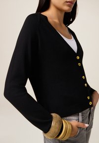 Schwarzer Cardigan aus weichem Strickmaterial, mit tiefem V-Ausschnitt und goldenen Knöpfen, kombiniert mit grauen Hosen und gestapelten Armbändern.