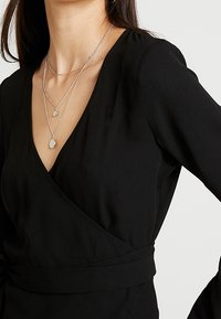 Sort omslagsbluse med dyb V-udskæring og lange ærmer. Har en tætsiddende talje og tre lagdelte sølvhalskæder med vedhæng.