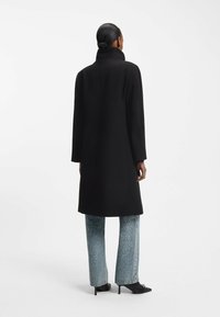 Zwarte wollen jas met een hoge kraag, op maat gemaakte silhouet en lange mouwen. Draagt het over lichtblauwe jeans met zilveren versieringen en zwarte hakken.