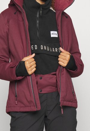 Person iført en bordeaux jakke over en sort halv-zip top med hvid skrift, der åbner jakken for at vise designet på trøjen.