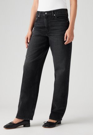 Žena v černých džínech s vysokým pasem a rovnými nohavicemi, černých balerínkách a bílém topu bez rukávů, stojící před jednobarevným pozadím.