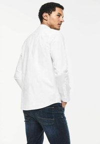 Biała, długasleeve koszula z kołnierzykiem zapinanym na guziki, pokazana z tyłu. Wykonana z gładkiego materiału, zestawiona z ciemnoniebieskimi dżinsami.