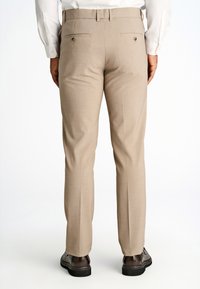 Pantaloni beige su misura con una texture liscia, taglio dritto e due tasche posteriori con chiusura a bottone. Indossati con scarpe eleganti nere.