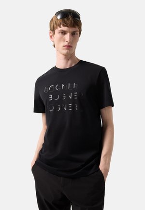 Młody mężczyzna z krótkimi włosami, noszący na głowie czarne okulary przeciwsłoneczne, czarną koszulkę z napisem "BOGNER" oraz czarne spodnie, ręce w kieszeniach.