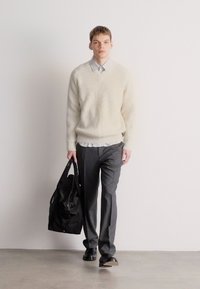 Ανοιχτό μπεζ ριμπ πουλόβερ πάνω από πουκάμισο με γιακά, σκούρες γκρι παντελόνες και μαύρη τσάντα. Το μοντέλο στέκεται μπροστά σε λευκό φόντο.