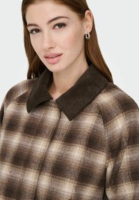 Ung kvinne med lys hud og brunt hår, iført sølvfargede creol-øreringer og en brun rutete frakk med mørkribbete krage, vendt mot kameraet.