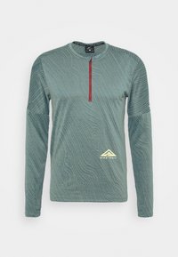 Long-sleeve teal top with textured design, half-zip front, and red zipper. Features a yellow logo on the lower left.