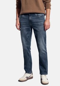 Homme portant un jean bleu, un sweat-shirt marron et des baskets blanches, se tenant avec les bras détendus le long du corps devant un fond uni.