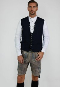 Marineblaue Wollweste mit silbernen Knöpfen, kombiniert mit grauen Leder-Shorts mit blauer floralem Stickerei und gelben Nähten, knielange schwarze Socken.