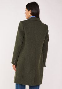 Cappotto in misto lana verde con un design strutturato, stile monopetto e accenti a tre bottoni sulle maniche. Collo piatto e spacco posteriore.
