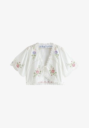 Jonge vrouw met zonnebril draagt een witte bloemen crop-top, witte rok, bruine kniehoge laarzen en armbanden, staat op grind vlakbij groen en een huis.