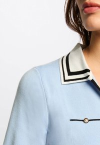 Helles blaues Hemd mit Kragen und kontrastierendem schwarzen Besatz; zeigt eine kleine Perle als Akzent und gerippte Textur. Nahaufnahme des Kragens und des oberen Oberteils.