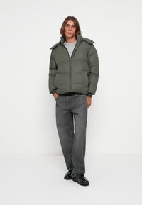 Padded olive green jacket with a stand-up collar, front zipper, and side pockets, paired with gray jeans and black shoes.