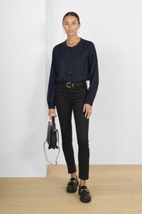 Camisa de botones azul marino con mangas abullonadas, combinada con jeans negros ajustados y loafers negros gruesos. Sosteniendo un pequeño bolso negro.