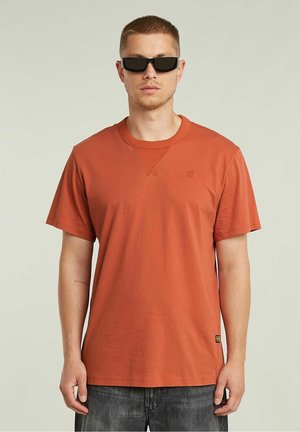 T-shirt a maniche corte di colore ruggine, realizzato in morbido cotone, con scollatura a girocollo, logo discreto e vestibilità rilassata, abbinato a jeans scuri.