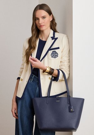 Žena s dugom kosom nosi krem sako s tamnoplavim obrubom, tamnoplavu majicu, široke traperice i zlatnu narukvicu, držeći veliku tamnoplav u kožu torbu za nošenje.