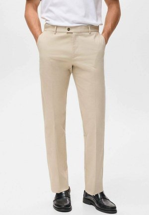 Beige Hose mit geradem Schnitt, aus glattem Stoff gefertigt, mit eingeprähten Falten. Kombiniert mit schwarzen Schuhen für einen Kontrast.