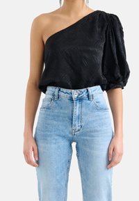 Schwarzes Top mit einem Schulterträger aus strukturiertem Stoff und puffigem Ärmel, kombiniert mit hochgeschnittenen, hellblauen Jeans.