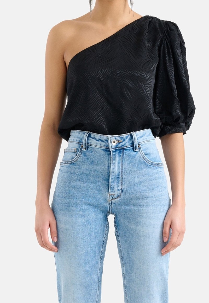 Schwarzes Top mit einem Schulterträger aus strukturiertem Stoff und puffigem Ärmel, kombiniert mit hochgeschnittenen, hellblauen Jeans.