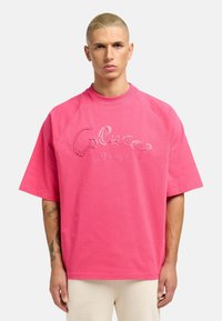 Lyserød bomulds oversized t-shirt med korte raglanærmer, prydet med broderet tekst "Colucci la Famiglia" på forsiden. Glat tekstur.