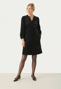 Robe en velours côtelé noir avec un décolleté en V, des manches bouffantes et des poches latérales. Texture lisse, longueur genoux, assortie à des chaussures noires à bout pointu.