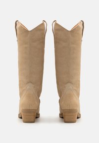 Botas de ante beige hasta la rodilla con punta afilada, pequeñas pestañas en la parte trasera y un tacón apilado. Presenta una textura suave y minimalistas. 