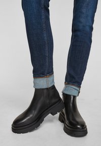 Black leather ankle boots with a thick rubber sole, featuring a zipped side. Paired with dark blue jeans rolled at the cuffs.