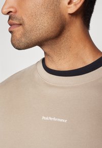 Beige színű pulóver kerek nyakkal, sötétkék belső gallérral és fehér "Peak Performance" logóval az elején. Puha tapintás.