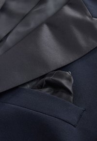 Veston de costume bleu marine avec revers en satin, présentant un mouchoir de poche en tissu sombre et transparent dépassant de la poche poitrine. Mélange de laine texturée.