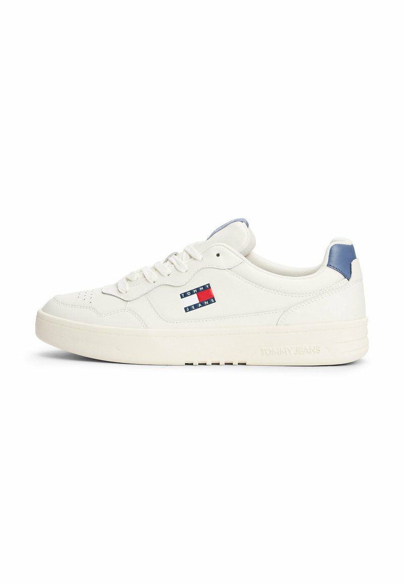 Zapatilla de cuero blanca con punta redondeada, suela de goma plana, acento azul en el talón y un logo de Tommy Jeans en rojo, blanco y azul.