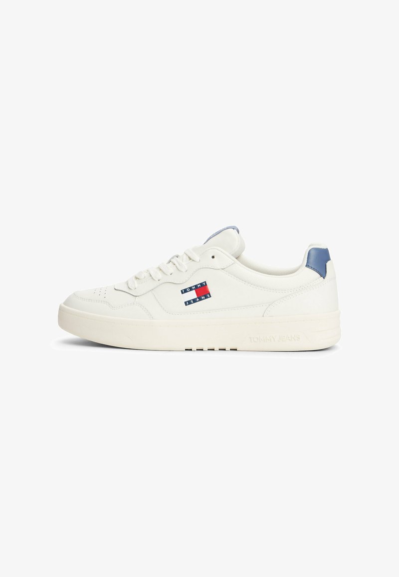 Zapatilla de cuero blanca con punta redondeada, suela de goma plana, acento azul en el talón y un logo de Tommy Jeans en rojo, blanco y azul.